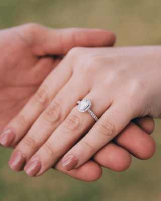 A close-up of two hands: one person holding another’s hand, showcasing an engagement ring on the ring finger. The background is blurred green.