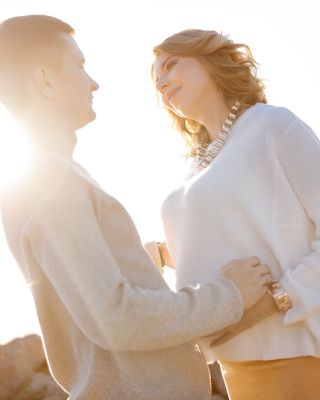A couple is standing outdoors near rocks, with the sun shining brightly behind them, appearing to enjoy a peaceful moment together.