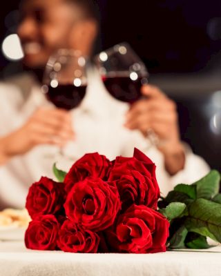 A couple enjoys a romantic dinner, toasting with wine glasses. Red roses and candles are on the table, adding to the intimate atmosphere.