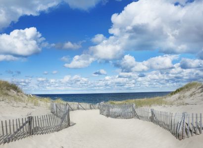 A sandy path with wooden fencing leads through dunes toward a calm ocean under a bright blue sky with fluffy white clouds ending the sentence.