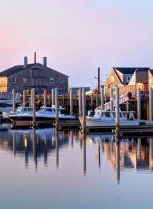 The image shows a peaceful harbor at dawn or dusk, featuring several boats docked by wooden buildings, with their reflections in the calm water.