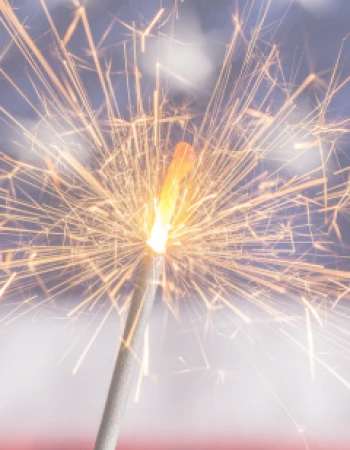 A close-up of a lit sparkler with bright sparks, set against a blurred background resembling the United States flag.