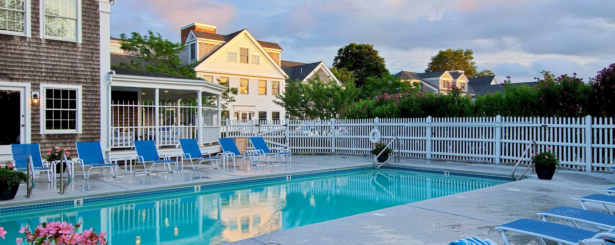 The image shows a serene outdoor swimming pool surrounded by blue lounge chairs, white fencing, and a charming backdrop of residential homes.