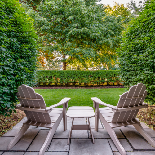 Two Adirondack chairs with footrests and a small table are placed on a stone patio, overlooking a lush garden with a large tree.