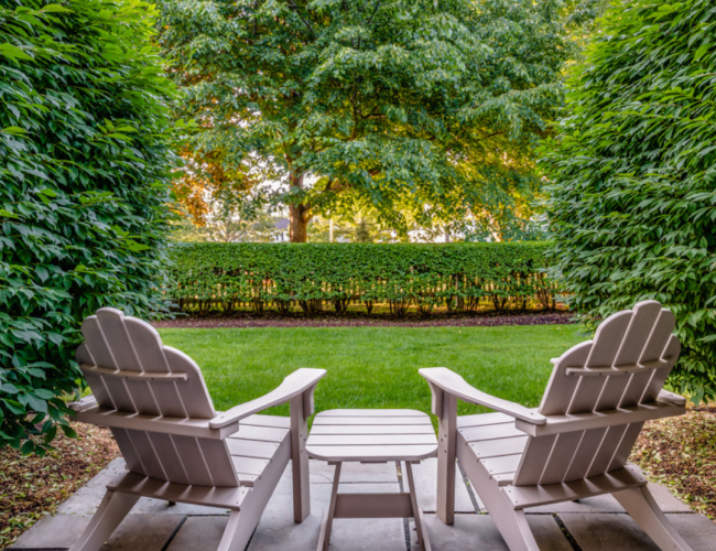 Two Adirondack chairs with footrests and a small table are placed on a stone patio, overlooking a lush garden with a large tree.