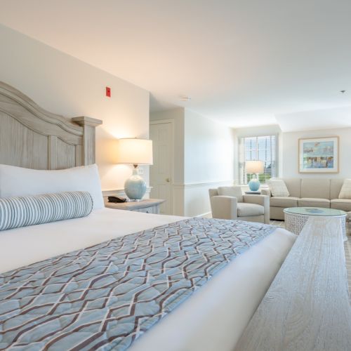 A bright and spacious hotel room featuring a large bed, a cozy seating area with sofas, and a light, airy decor with soft blue accents.
