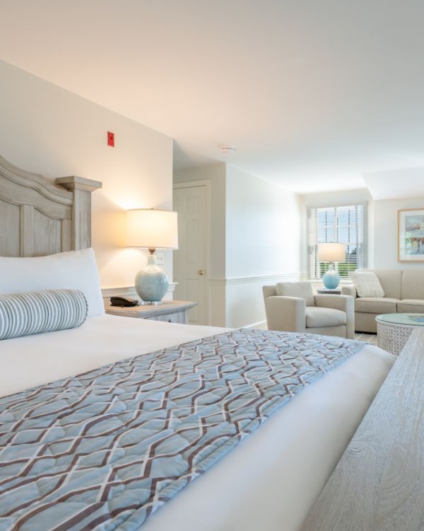 A bright and spacious hotel room featuring a large bed, a cozy seating area with sofas, and a light, airy decor with soft blue accents.